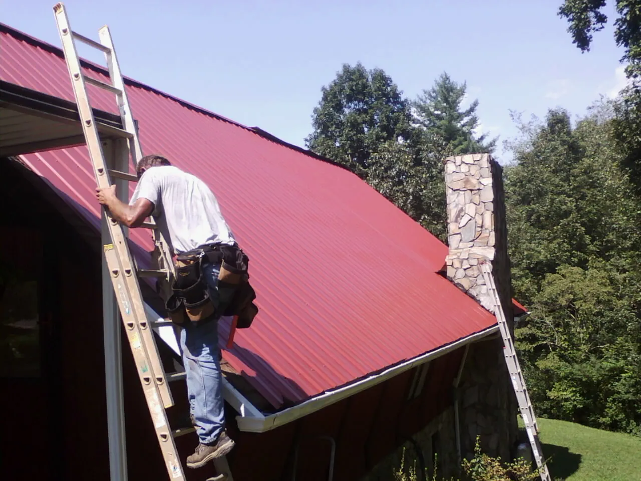Roofer on a ladder beside a red metal roof and stone chimney — Soffit & Fascia Repair by Roofing Brothers in Casper